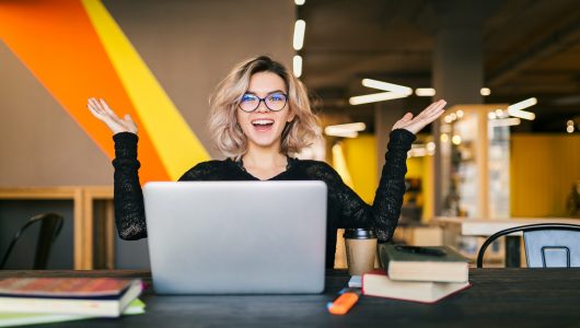 funny happy excited young pretty woman sitting at table in black shirt working on laptop in co-working office, wearing glasses